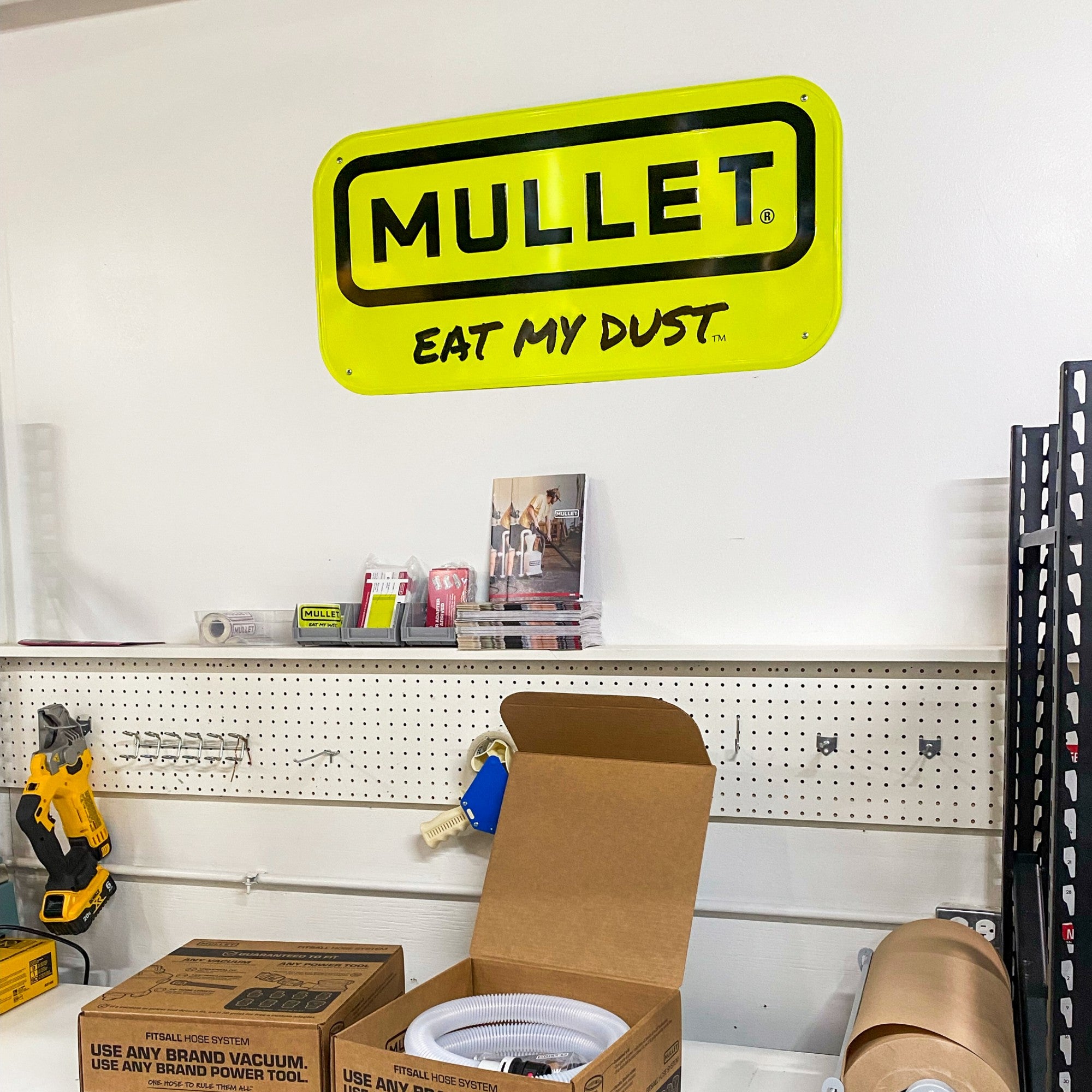 Yellow 'Mullet Eat My Dust' sign on a white wall with tools and boxes below.
